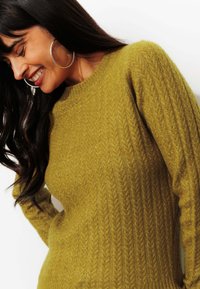Pull en tricot vert olive avec un motif texturé et des poignets en côtes. Encolure ronde et manches longues, matériau doux.