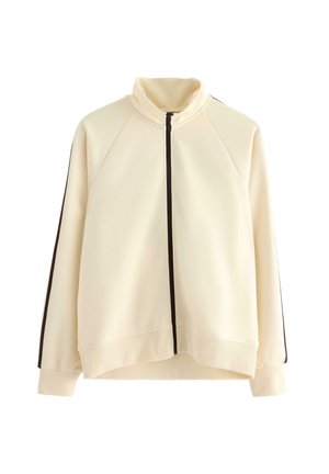 Krémová zipová bunda s vysokým límcem, dlouhými rukávy a tmavými pruhy běžícími po každém rukávu.