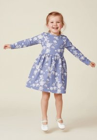 Fioletowa sukienka w kwiaty z długimi rękawami, okrągłym dekoltem i marszczonym pasem. Posiada białe wzory kwiatowe i rozkloszowany dół. Noszona z białymi butami.