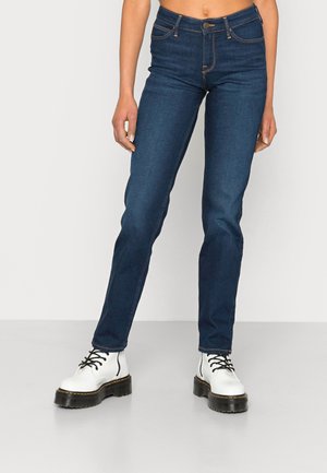 Personne portant un jean skinny bleu foncé et des bottes blanches à lacets avec des semelles épaisses noires, debout sur un sol gris clair devant un fond uni.