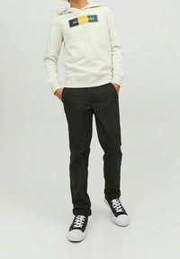 Person klädd i en ljusgrå hoodie med "Jack & Jones"-logotyp, svarta byxor och svart-vita sneakers, stående med händerna i fickorna.