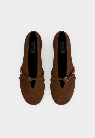 Καφέ σουέντ μπαλαρίνες με ένα μικρό λουράκι με αγκράφα και χρυσό μενταγιόν με το λογότυπο Moschino, σε θέα από πάνω σε λευκό φόντο.