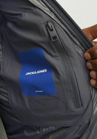 Interiér šedej bundy so zapínacím vreckom, hladkou textúrou a modrou nálepkou s nápismi "JACK & JONES" a "ORIGINALS" v bielej farbe.