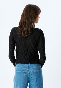 Schwarzes Langarmoberteil mit strukturierter Blattform, semi-transparenter Stoff, figurbetonter Schnitt, kombiniert mit hochgeschnittenen blauen Jeans.