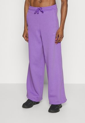 Hombre con pantalones deportivos Adidas morados y holgados con cintura de cordón y zapatos negros sin cordones, de pie sobre un fondo blanco liso.