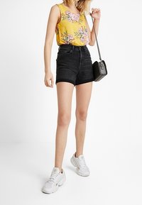 Gulblommig linne i kombination med högmidjade svarta denimshorts; modellen bär vita sneakers och håller en svart axelremsväska.