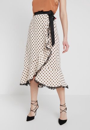 Femme portant une jupe midi portefeuille blanche à pois marron, bordée de dentelle noire, nouée avec une ceinture noire, assortie à des chaussures noires à talons hauts et lacets.