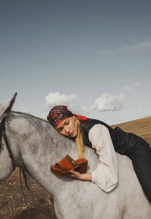 Femme portant un foulard à motifs allongée sur un cheval gris, tenant une sandale compensée marron, dans un décor extérieur désertique sous un ciel nuageux.