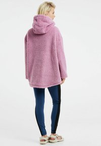 Felpa in pile morbido e rosa, oversize, con cappuccio, indossata su leggins blu con strisce nere ai lati e sandali chiari.