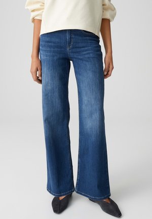 Blaue High-Waist-Jeans mit weitem Bein, getragen mit schwarzen spitzen Schuhen und einem cremefarbenen Langarmshirt, gezeigt vom mittleren Oberkörper bis zu den Füßen.