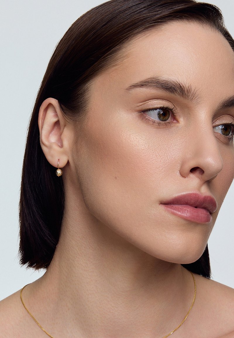 Złoty kolczyk w formie obręczy z teksturowanym okrągłym detalem, noszony na uchu modelki. Modelka ma gładką skórę i proste, ciemne włosy.