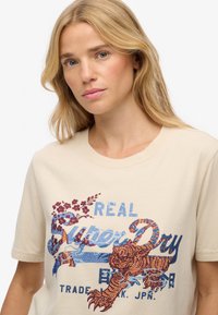 Kremowa koszulka z krótkim rękawem z niebieskim i pomarańczowym nadrukiem graficznym, przedstawiająca tygrysa oraz motywy florystyczne, wykonana z miękkiego materiału bawełnianego.