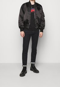 Chaqueta bomber de satén negra con puños acanalados, cierre frontal de cremallera y bolsillos laterales. Combinada con jeans negros y botas negras con cordones.