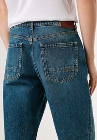 Blå denim jeans med klassisk snit, to baglommer med syninger og et brunt læderbæltemærke.