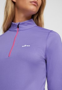 Lila sportliches Langarmshirt mit hohem Kragen und einem halben Reißverschluss. Mit pinkem Reißverschlussdetail und weißem Logo auf der Brust. Glatte Textur.