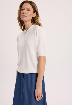 Kobieta o jasnobrązowych włosach, ubrana w białą koszulkę polo z krótkim rękawem i niebieską spódnicę, stojąca na tle jednolitego, jasnego tła.