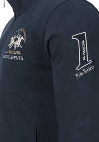 Marinblå sweatshirt med hög krage, med broderad hästgrafik och text, varmt tyg och sidoficka med dragkedja.