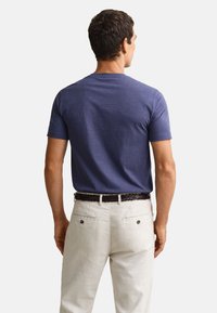 Mand iført en ensfarvet blå t-shirt med korte ærmer, stoppet ned i lyse beige bukser med et mørkt vævet bælte, set bagfra.