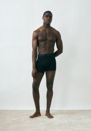 Lihaseline mees seisab paljajalu vaibal, kandes musta Calvin Kleini bokseritekkide püksikuid, valge ühevärvilise tausta taustal.