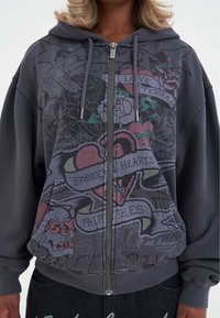 Felpa con zip grigia scura caratterizzata da intricati disegni grafici con rose, cuori e testo. Realizzata in un tessuto morbido, dispone di tasche frontali e coulisse.