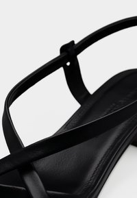 Μαύρο δερμάτινο σανδάλι με λεπτούς σταυρωτούς ιμάντες και χαμηλό τετράγωνο τακούνι σε λευκό φόντο.