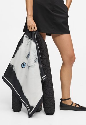 Czarna pikowana kurtka trzymana w jednej ręce, z białym szalikiem z grafiką kota, niebieskimi oczami i czarną obwódką. Na nogach czarne płaskie buty.