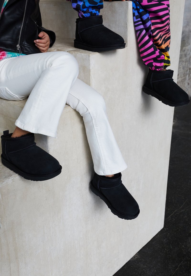 Deux personnes portant des bottines noires sont assises sur des marches en béton, l'une en pantalon blanc, l'autre en pantalon à motifs colorés.