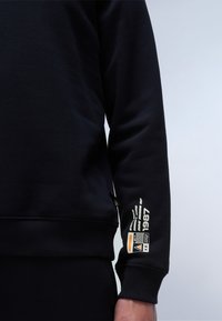 Sweat à capuche noir avec des poignets et un ourlet côtelés. Présente un texte imprimé et un design graphique sur la manche. Fabriqué en matériau doux et extensible.