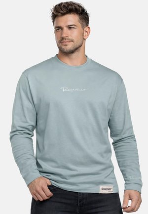 Mann mit kurzen Haaren, der ein hellblaues Langarmshirt mit weißem Schriftzug auf der Brust und einem Etikett nahe dem Saum trägt, posiert vor einfachem Hintergrund.