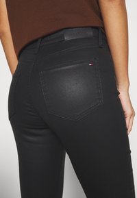 Jeans en denim noir avec une texture lisse et des détails cousus. Présente une poche arrière avec une petite étiquette de logo rouge, blanc et bleu.
