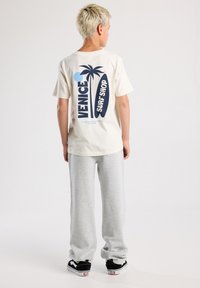 Cream T-shirt featuring blue and dark blue "VENICE SURF SHOP" graphic and palm tree design on the back, paired with gray sweatpants and black sneakers.