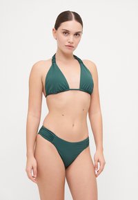Tumši zaļš bikini komplekts, kas sastāv no trīsstūrveida haltera topa un atbilstošiem zema vidukļa apakšdelmiem ar savilkuma detaļām sānos. Gluda tekstūra.