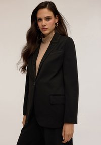 Blazer nero con un bottone singolo, revers a raglan e taschino sul petto. Presenta dettagli in cucitura marrone ed è realizzato in un tessuto liscio.
