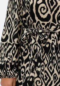 Robe noire et beige à motifs avec un tissu texturé, dotée de manches longues et d'une taille nouée. Le motif comprend des volutes et des formes en losange.