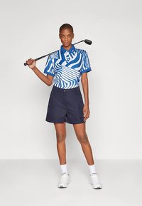 Rövid ujjú kék póló fehér absztrakt mintákkal, tengerészkék rövidnadrággal párosítva. A modell golfütőt tart, fehér cipőt és zoknit visel.