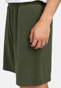 Green cotton shorts with an elastic waistband, featuring side pockets and a logo embossed at the hem. Soft texture and relaxed fit.