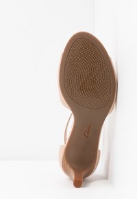 Sandale à talon bas, dessus beige et semelle en caoutchouc texturée affichant le logo "Clarks", positionnée debout contre un coin blanc.