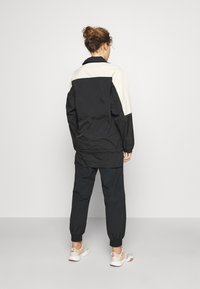 Svart och krämfärgad lång jacka i lätt material, med hög krage och elastiska ärmslut, kombinerad med svarta smalrandiga byxor.