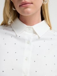 Hvid skjorte med knaplukning, der har en spids krave, prydet med små firkantede sølvpalietter arrangeret i et spredt mønster over stoffet.