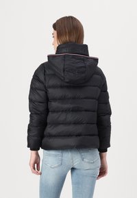 Svart pufferjakke med hette og horisontal quilted design. Utsmykket med en rød og hvit stripete krage og logo tekst bak.