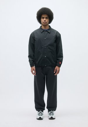 Pánska čierna, ľahká bunda s gombíkovým zapínaním, elastickými manžetami a vreckami, v kombinácii s tmavými nohavicami a bielymi teniskami.