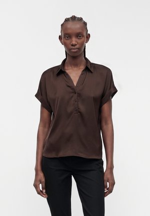Femme aux cheveux tressés portant un chemisier marron foncé à manches courtes boutonné et un pantalon noir, debout devant un fond clair uni.