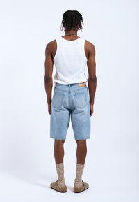 Lichtblauwe denim shorts met een relaxte pasvorm, voorzien van twee achterzakken en een bruine label. Gecombineerd met een witte tanktop en beige schoeisel.
