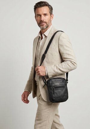 Mand i beige jakkesæt bærer en sort læder crossbody-taske på skulderen mod en ensartet lys baggrund.