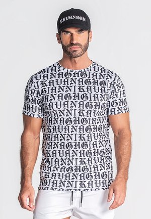 Uomo con cappellino nero e T-shirt bianca coperta da testo nero ripetuto "KAVANAGH", abbinata a pantaloncini bianchi, su sfondo uniforme.