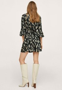 Robe noire courte avec un motif de feuilles, taille ceinturée et manches évasées. Portée avec des bottes blanches hautes et un talon empilé marron.