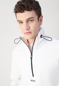 Hvid halv-zip pullover lavet af fleece med sort lynlås og hardware-detaljer. Har et diskret logo på brystet og strukturerede sidestykker.