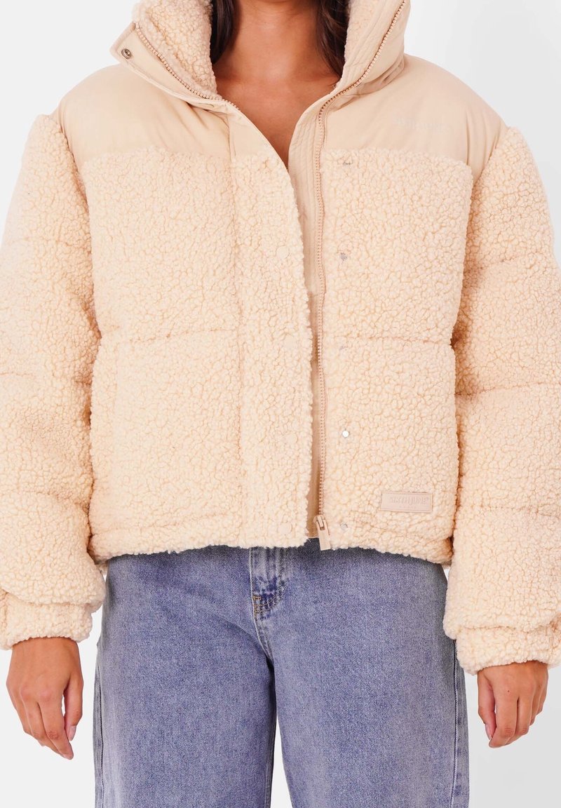 Personne portant une veste en polaire sherpa beige avec une capuche et un jean en denim bleu clair, vue des épaules jusqu'aux hanches.
