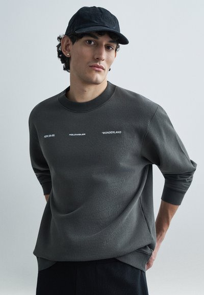 Donkergrijze sweatshirt met een ronde hals, voorzien van witte tekstgrafieken. Stof heeft een gladde textuur, met iets verlaagde schouders en geribbelde manchetten.