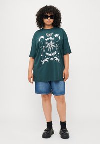 T-shirt oversize verde scuro con stampa grafica bianca, che presenta una farfalla, una palma e del testo. Abbinato a pantaloni shorts di jeans e scarpe nere chunky.